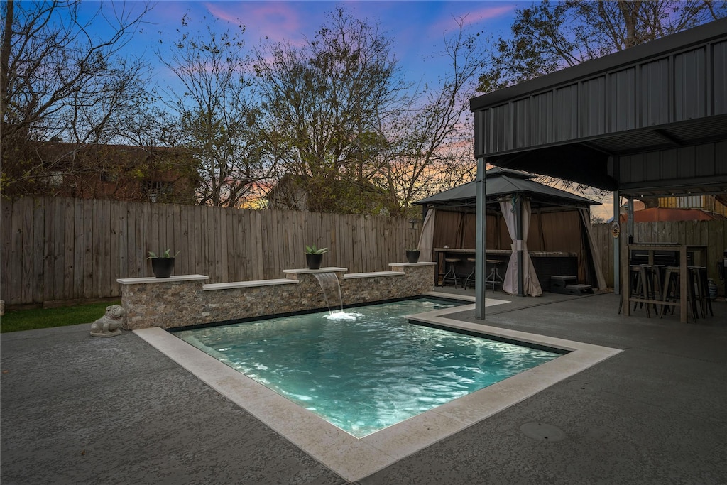 pool at dusk with pool water feature, a gazebo, a patio, and exterior bar