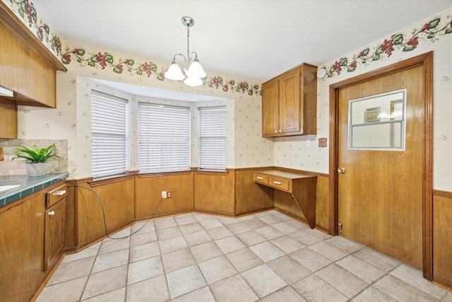 kitchen featuring brown cabinets, a wainscoted wall, tile countertops, a chandelier, and wallpapered walls