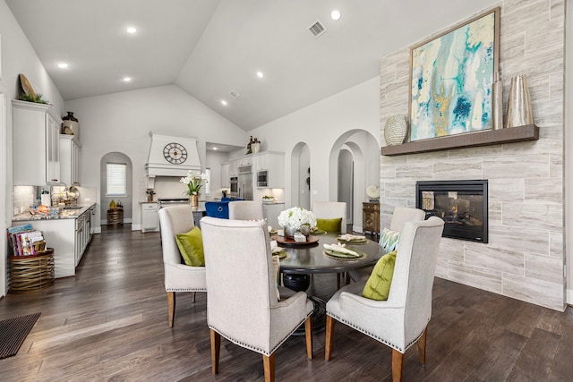 dining area with arched walkways, high vaulted ceiling, dark wood-style flooring, and a tiled fireplace