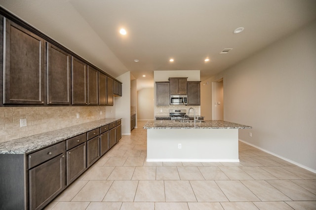 kitchen with light stone countertops, stainless steel appliances, light tile patterned flooring, a center island with sink, and dark brown cabinets