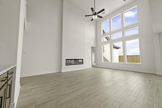 26515 Castra Crescent Trl, Richmond, TX 77406 - unfurnished living room featuring a towering ceiling, light wood-style flooring, ceiling fan, a glass covered fireplace, and baseboards