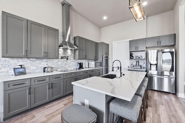 kitchen featuring wall chimney range hood, stainless steel appliances, a center island with sink, and gray cabinets