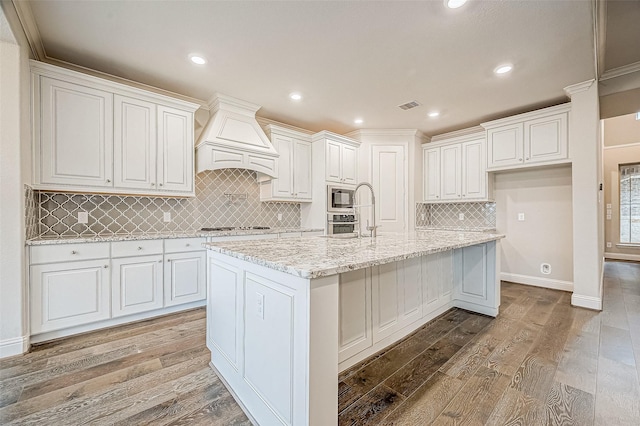 kitchen featuring custom exhaust hood, white cabinetry, stainless steel appliances, a kitchen island with sink, and crown molding