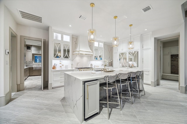 kitchen featuring a kitchen island with sink, decorative light fixtures, custom range hood, and white cabinets