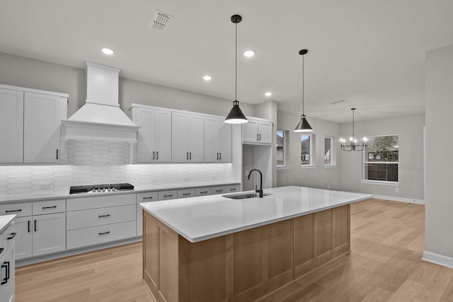 kitchen with sink, a kitchen island with sink, backsplash, custom range hood, and stainless steel gas stovetop