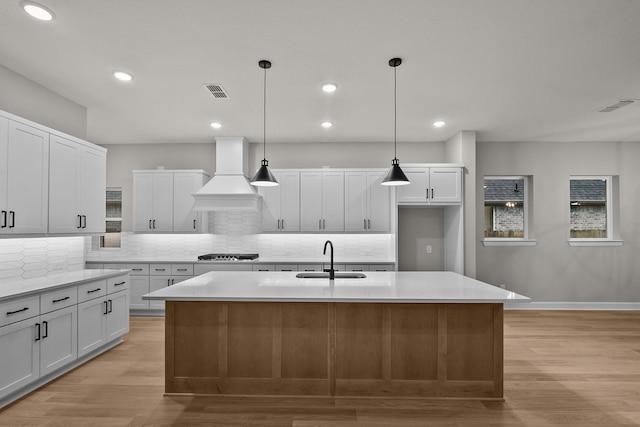 kitchen featuring premium range hood, sink, white cabinetry, hanging light fixtures, and an island with sink