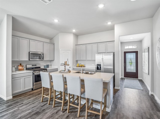 kitchen featuring stainless steel appliances, a sink, gray cabinets, dark wood finished floors, and a center island with sink