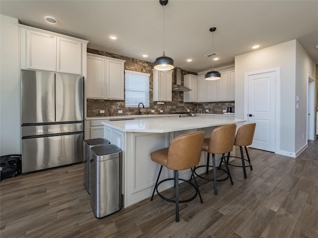 kitchen with wall chimney range hood, stainless steel refrigerator, hanging light fixtures, white cabinets, and a kitchen island