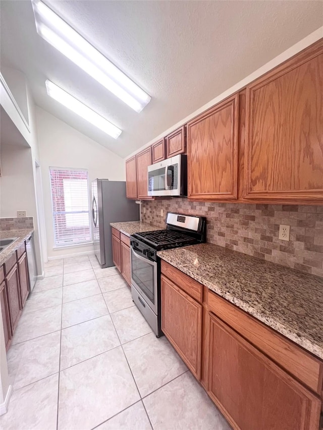 kitchen with appliances with stainless steel finishes, brown cabinets, light stone countertops, vaulted ceiling, and light tile patterned flooring