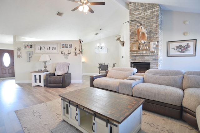 living room with vaulted ceiling with beams, ceiling fan with notable chandelier, a brick fireplace, and light wood-type flooring