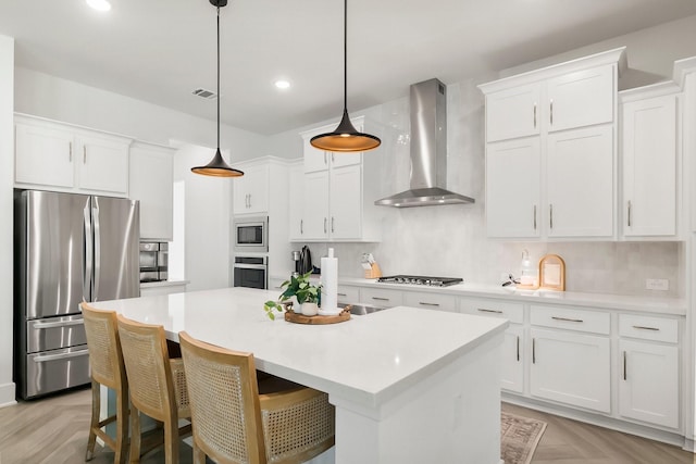 kitchen featuring white cabinetry, wall chimney range hood, an island with sink, stainless steel appliances, and light parquet floors