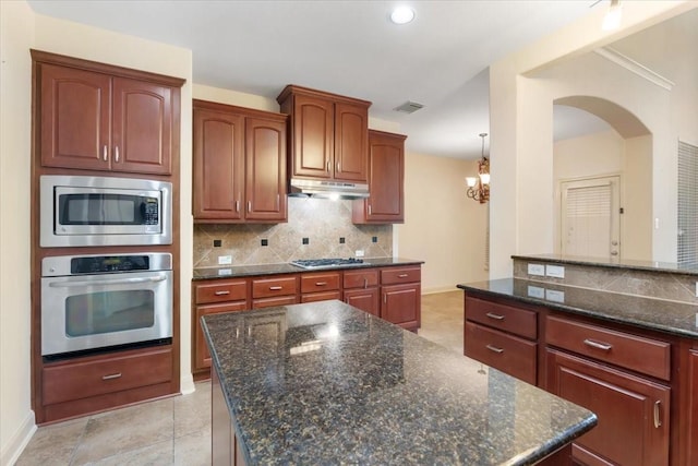 kitchen with appliances with stainless steel finishes, hanging light fixtures, backsplash, a center island, and dark stone counters