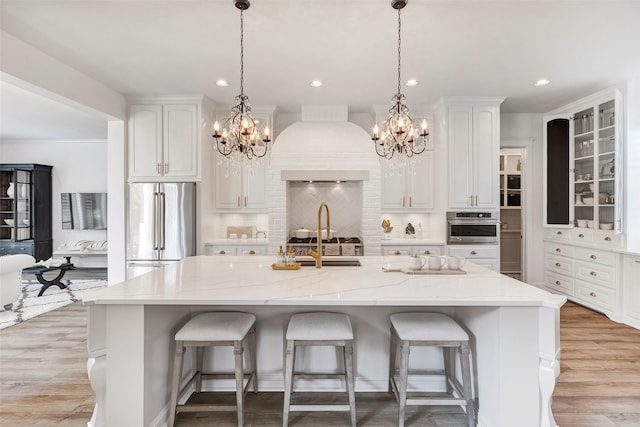 kitchen featuring premium range hood, stainless steel appliances, light stone counters, white cabinets, and a center island with sink