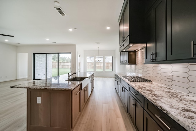 kitchen with tasteful backsplash, sink, stainless steel gas cooktop, light stone countertops, and a center island with sink