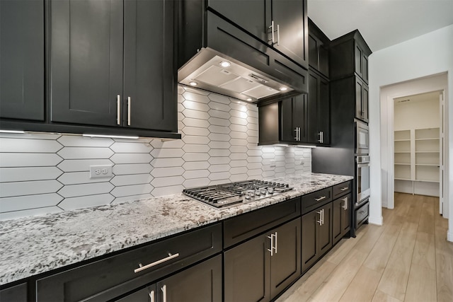 kitchen featuring appliances with stainless steel finishes, tasteful backsplash, light stone counters, light hardwood / wood-style floors, and exhaust hood