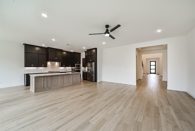 kitchen featuring light wood-type flooring, ceiling fan, stainless steel appliances, a kitchen island with sink, and backsplash