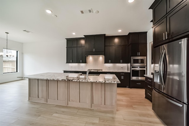 kitchen with light stone counters, light hardwood / wood-style flooring, pendant lighting, stainless steel appliances, and a kitchen island with sink