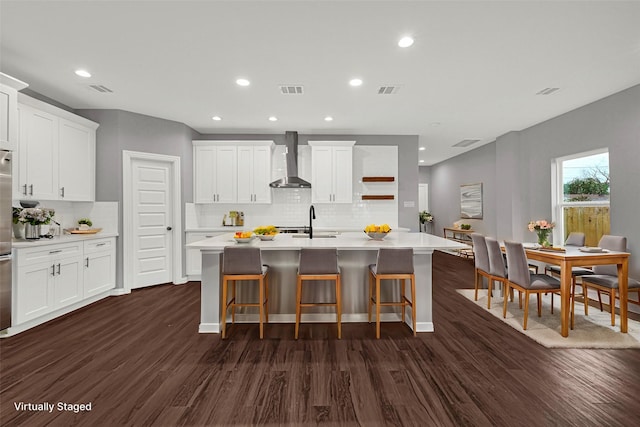 kitchen with light countertops, a center island with sink, wall chimney range hood, and white cabinetry