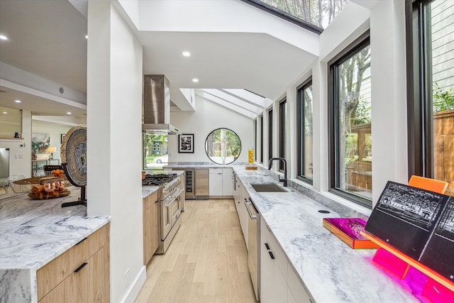 kitchen with wall chimney exhaust hood, sink, white cabinetry, light stone counters, and stainless steel appliances