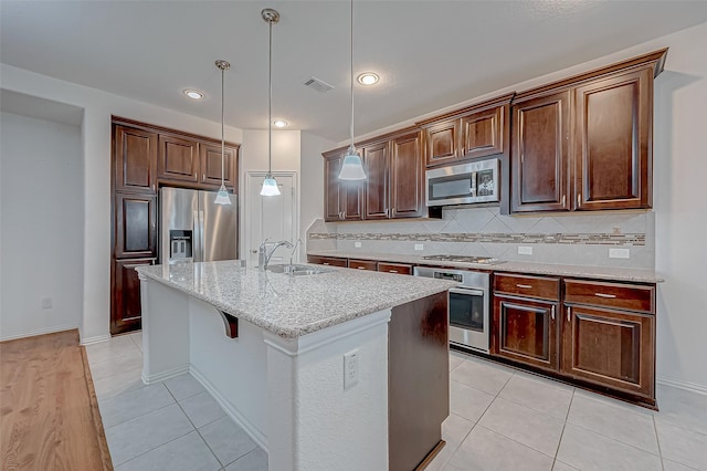 kitchen with pendant lighting, sink, a kitchen island with sink, stainless steel appliances, and light stone countertops