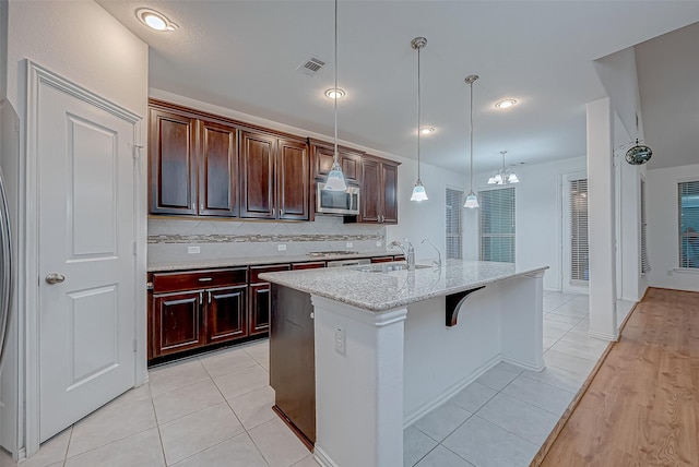 kitchen featuring sink, light tile patterned floors, light stone counters, a center island with sink, and decorative light fixtures