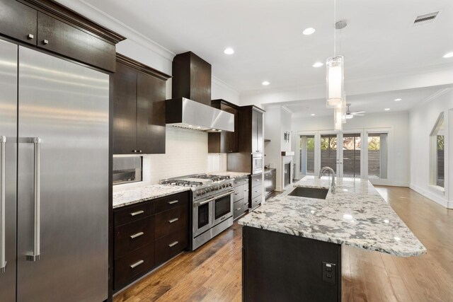 kitchen with wall chimney range hood, sink, hanging light fixtures, high quality appliances, and an island with sink