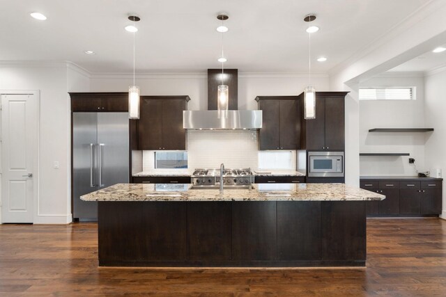 kitchen featuring decorative light fixtures, built in appliances, a kitchen island with sink, and wall chimney exhaust hood