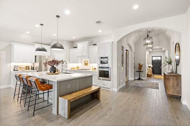 kitchen with sink, hanging light fixtures, stainless steel appliances, white cabinets, and a large island with sink