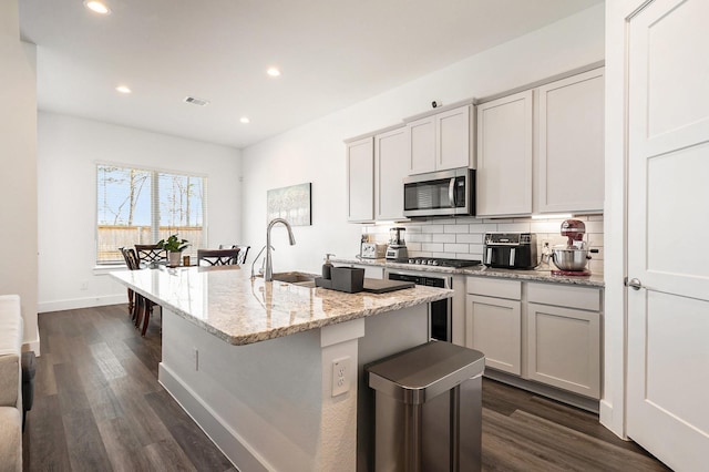 kitchen with appliances with stainless steel finishes, sink, dark hardwood / wood-style flooring, light stone countertops, and a center island with sink