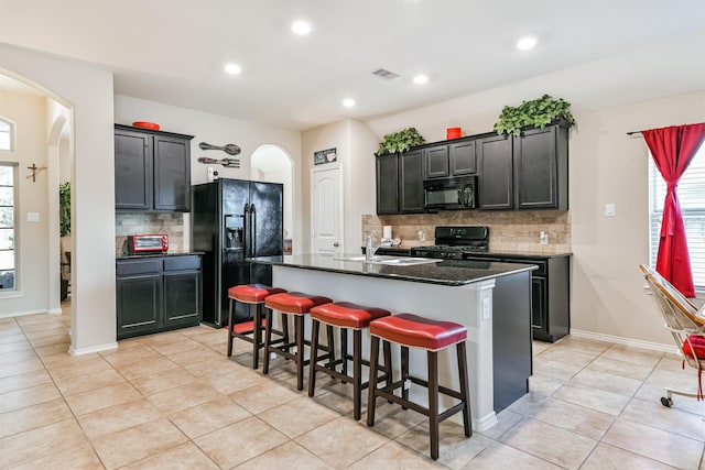 kitchen with sink, dark stone countertops, a center island with sink, black appliances, and light tile patterned flooring