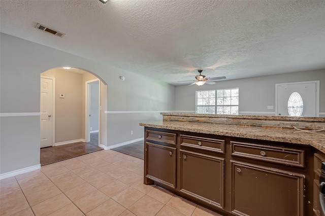 kitchen featuring dark brown cabinets, a textured ceiling, light tile patterned floors, ceiling fan, and light stone countertops