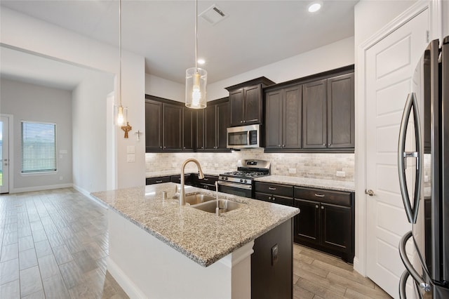 kitchen featuring sink, hanging light fixtures, stainless steel appliances, light stone countertops, and a center island with sink