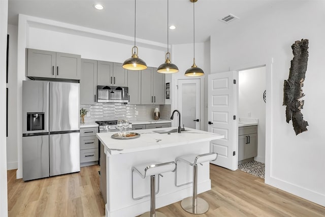 kitchen featuring decorative light fixtures, gray cabinetry, backsplash, a kitchen island with sink, and stainless steel appliances