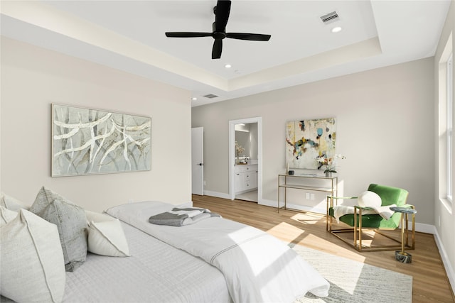 bedroom with ceiling fan, wood-type flooring, a tray ceiling, and ensuite bath