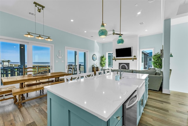 kitchen featuring a large fireplace, hanging light fixtures, stainless steel dishwasher, a center island with sink, and light wood-type flooring