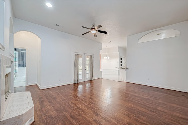 unfurnished living room with french doors, ceiling fan with notable chandelier, a tiled fireplace, and dark wood-type flooring