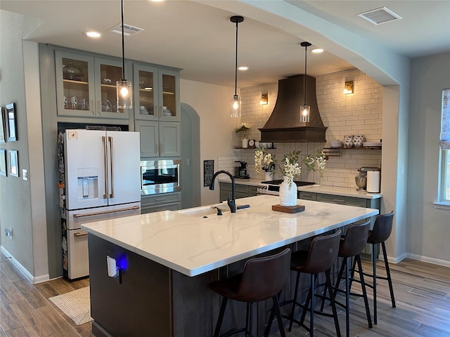 kitchen featuring appliances with stainless steel finishes, light stone countertops, custom range hood, an island with sink, and decorative backsplash