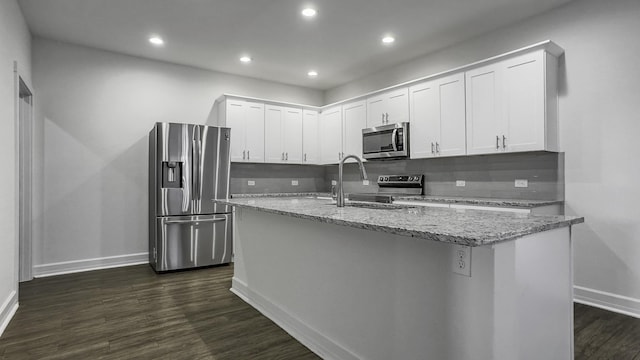 kitchen with white cabinets, dark hardwood / wood-style flooring, light stone counters, stainless steel appliances, and a center island with sink