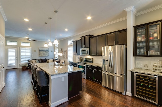 kitchen with appliances with stainless steel finishes, wine cooler, hanging light fixtures, a kitchen island with sink, and light stone counters