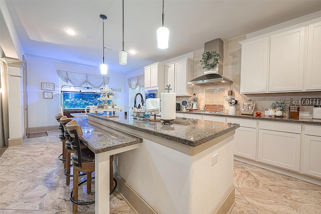 kitchen featuring a kitchen island with sink, hanging light fixtures, white cabinetry, and wall chimney exhaust hood