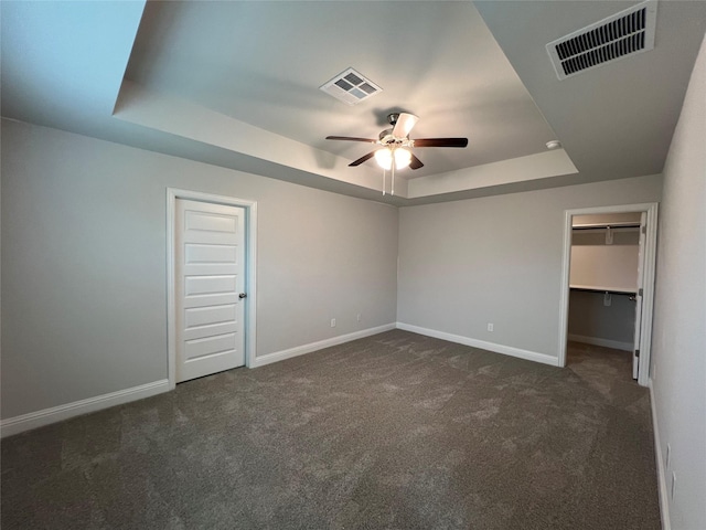 unfurnished bedroom featuring ceiling fan, a raised ceiling, dark carpet, a walk in closet, and a closet