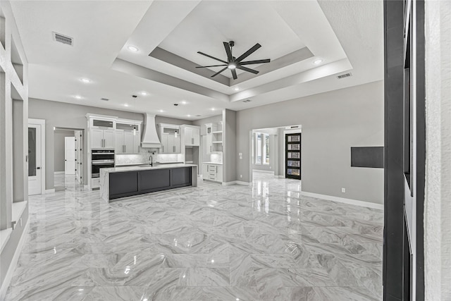 kitchen with sink, white cabinetry, custom range hood, an island with sink, and a raised ceiling