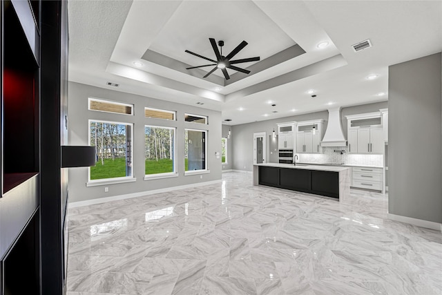 kitchen featuring sink, custom exhaust hood, white cabinetry, a center island with sink, and a tray ceiling