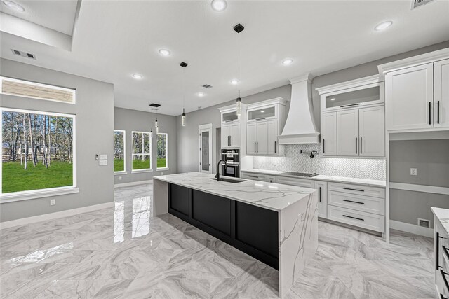 kitchen featuring pendant lighting, light stone countertops, an island with sink, custom range hood, and white cabinets