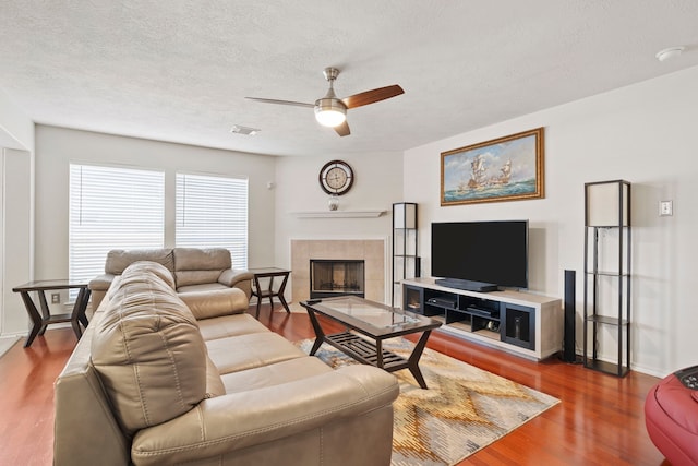 living room featuring a textured ceiling, wood-type flooring, a tile fireplace, and ceiling fan