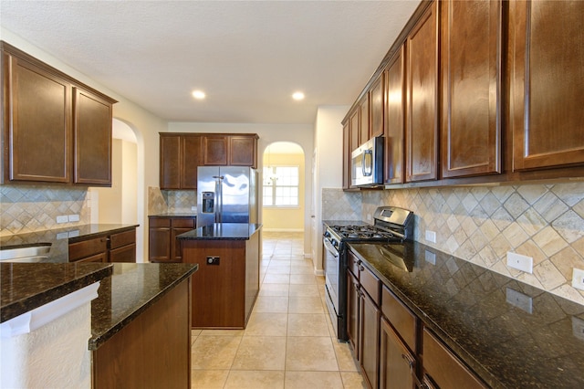 kitchen featuring light tile patterned flooring, tasteful backsplash, appliances with stainless steel finishes, a kitchen island, and dark stone counters