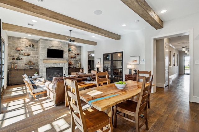 dining space with ceiling fan, dark hardwood / wood-style floors, beam ceiling, and a stone fireplace