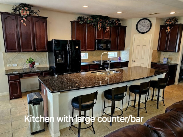 kitchen with an island with sink, light tile patterned floors, dark stone counters, and black appliances