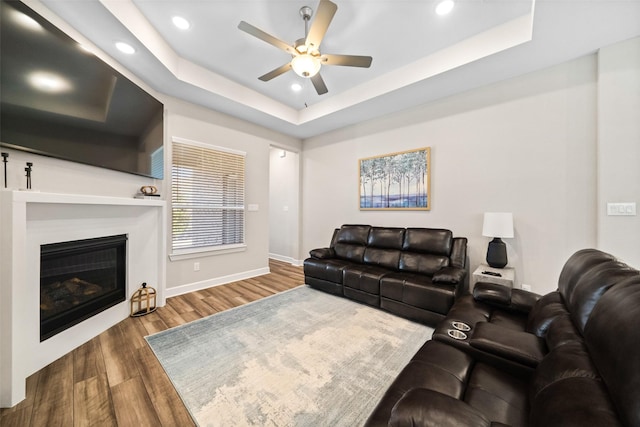 living room featuring wood finished floors, baseboards, a tray ceiling, ceiling fan, and a glass covered fireplace