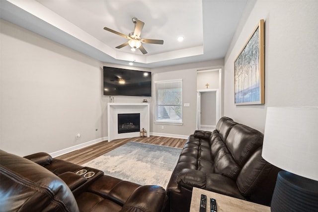 living area with a tray ceiling, wood finished floors, baseboards, and ceiling fan